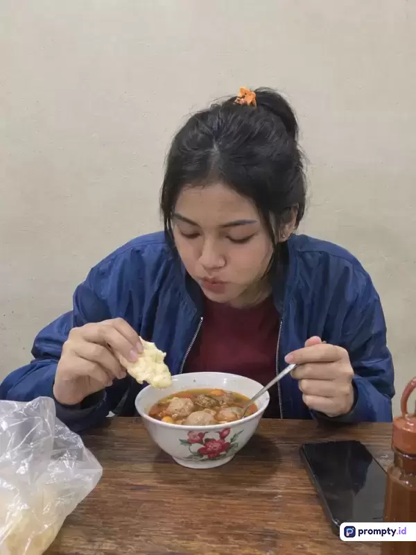 Gambar bergaya fotografi kasual ini menampilkan seorang wanita berambut hitam dengan gaya kuncir kuda mengenakan jaket biru tua sedang menikmati semangkuk bakso kuah hangat di meja kayu, dengan kata kunci kuliner, bakso, makan, wanita, dan indonesia, serta didominasi oleh warna biru tua, cokelat kayu, dan putih tulang.