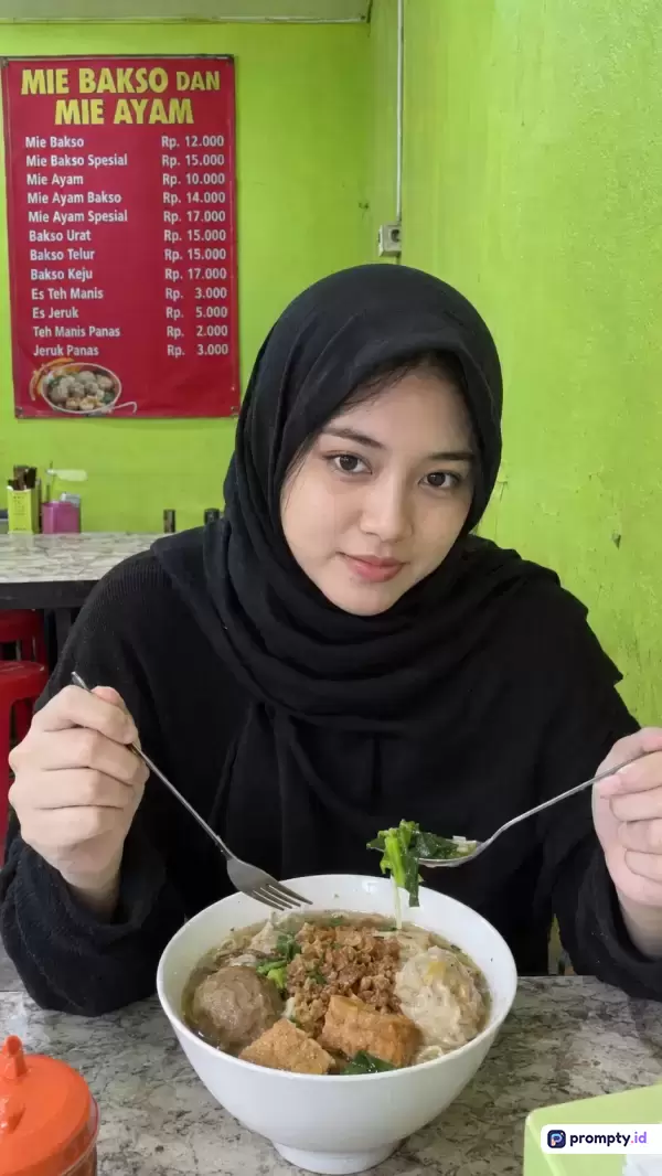 Foto bergaya kasual dan natural ini menampilkan seorang wanita berhijab hitam yang sedang tersenyum ramah sambil menikmati semangkuk bakso kuah hangat di sebuah warung sederhana dengan latar belakang dinding hijau cerah dan daftar menu merah, serta memiliki kata kunci kuliner, bakso, wanita berhijab, warung lokal, dan makanan Indonesia, dengan tiga warna dominan yaitu hijau, hitam, dan merah.