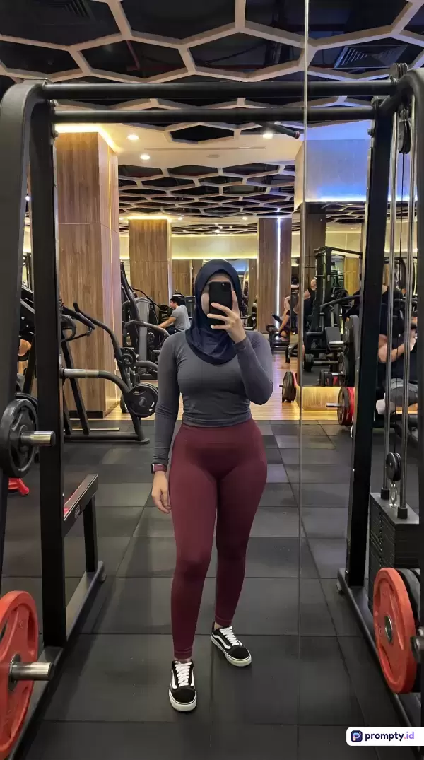 Foto bergaya swafoto cermin ini menampilkan seorang wanita berhijab yang sedang berpose di dalam gym modern dengan latar plafon bermotif sarang lebah dan peralatan kebugaran lengkap, di mana ia mengenakan pakaian olahraga ketat berlengan panjang abu-abu gelap dan celana legging merah marun, dengan kata kunci utama fitness, gym, wanita berhijab, olahraga, dan swafoto cermin, serta didominasi oleh palet warna abu-abu gelap, merah marun, dan hitam.